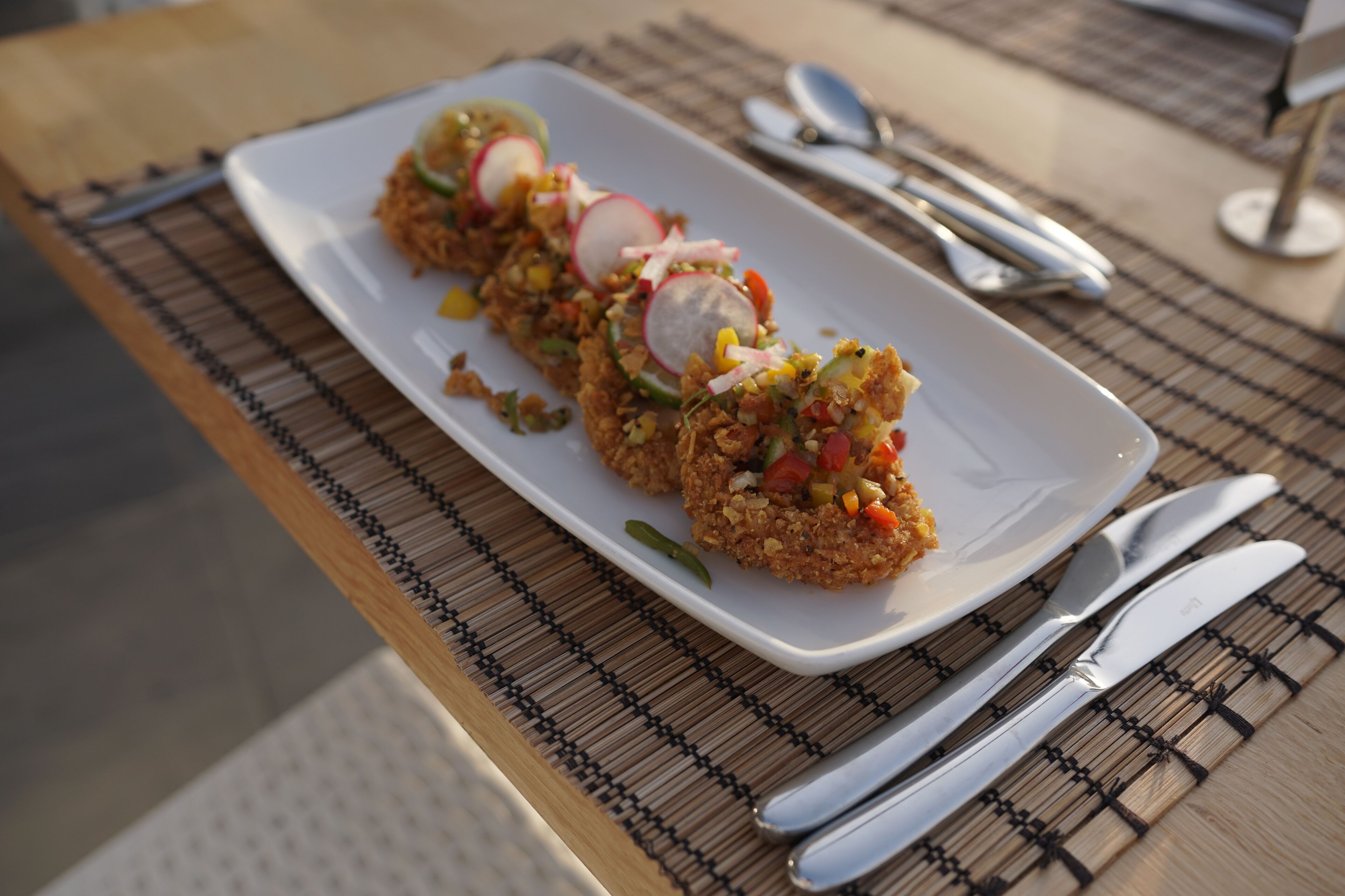 Une assiette rectangulaire blanche contenant un plat, accompagnée d’une fourchette et d’un couteau, le tout posé sur un tapis en bambou.