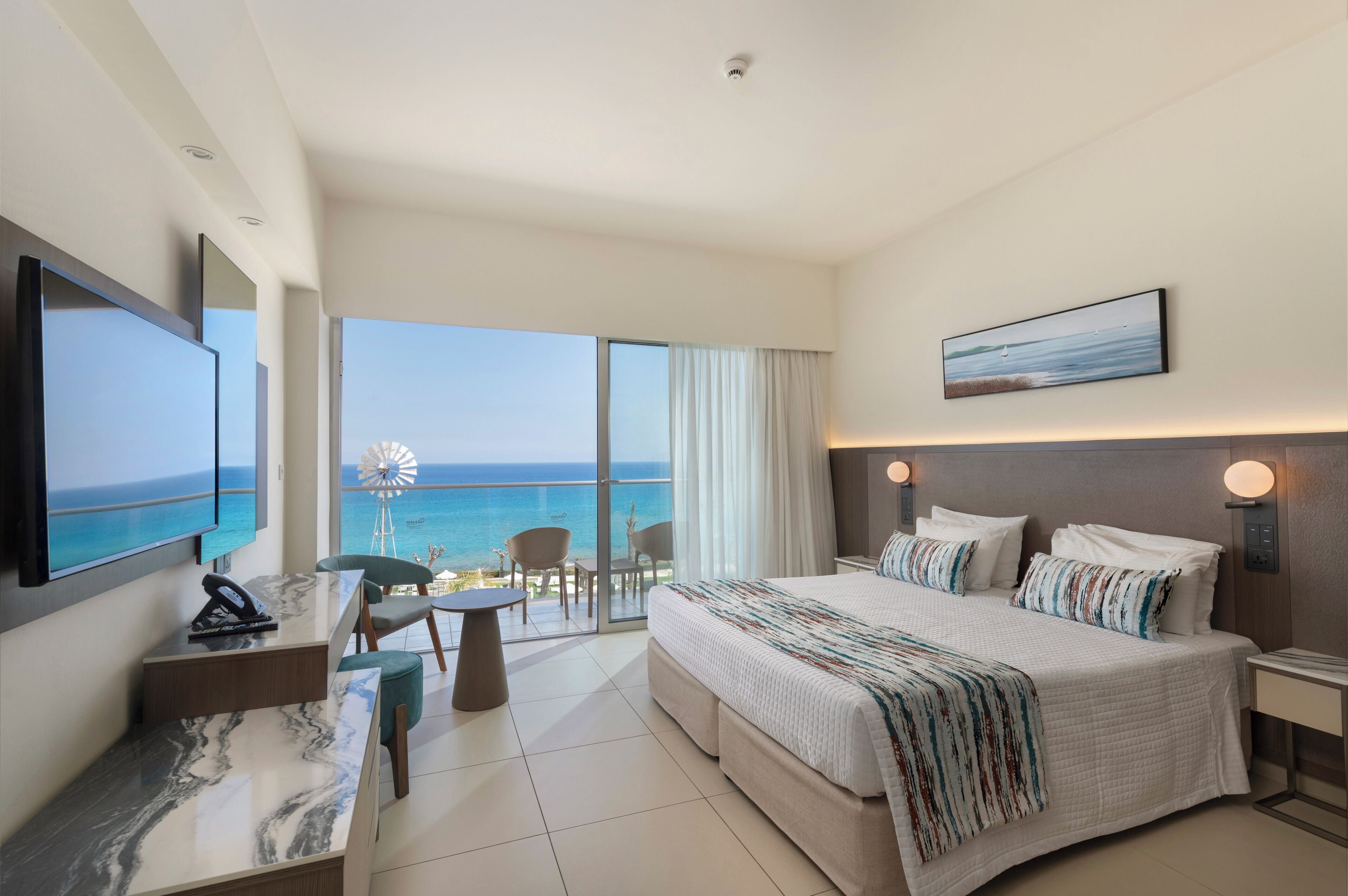 A hotel room with a large bed, a desk, a chair, a TV, and a view of the ocean.
