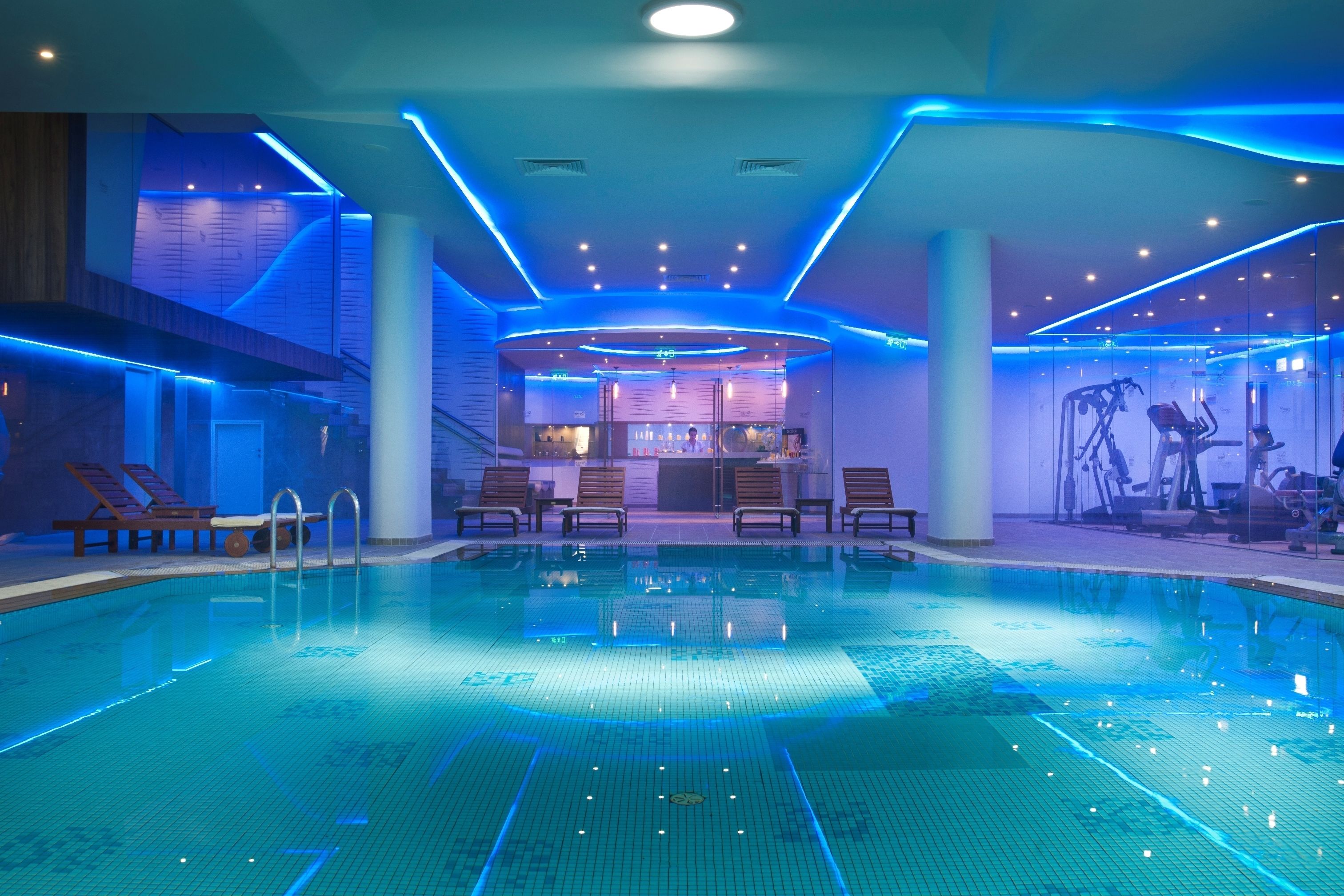 An indoor swimming pool with blue LED lighting, lounge chairs, and exercise equipment.