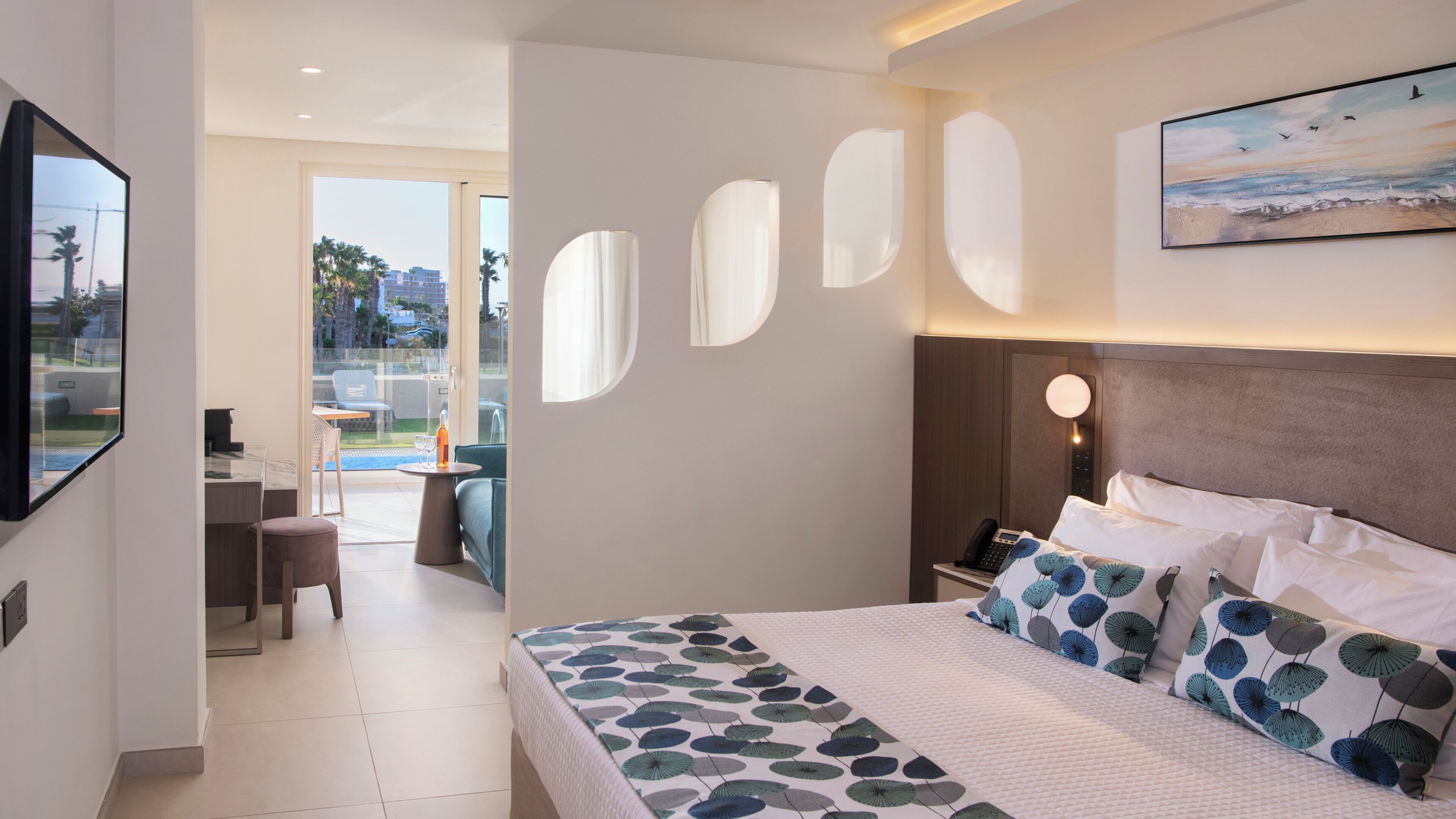 A hotel room with a bed, a desk, a chair, a balcony with a view, and a painting on the wall.
