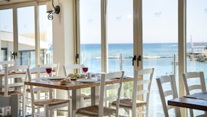 Un espace repas avec des chaises et des tables blanches, de grandes fenêtres offrant une vue sur la mer et les bateaux, et le logo Pernera sur les fenêtres.