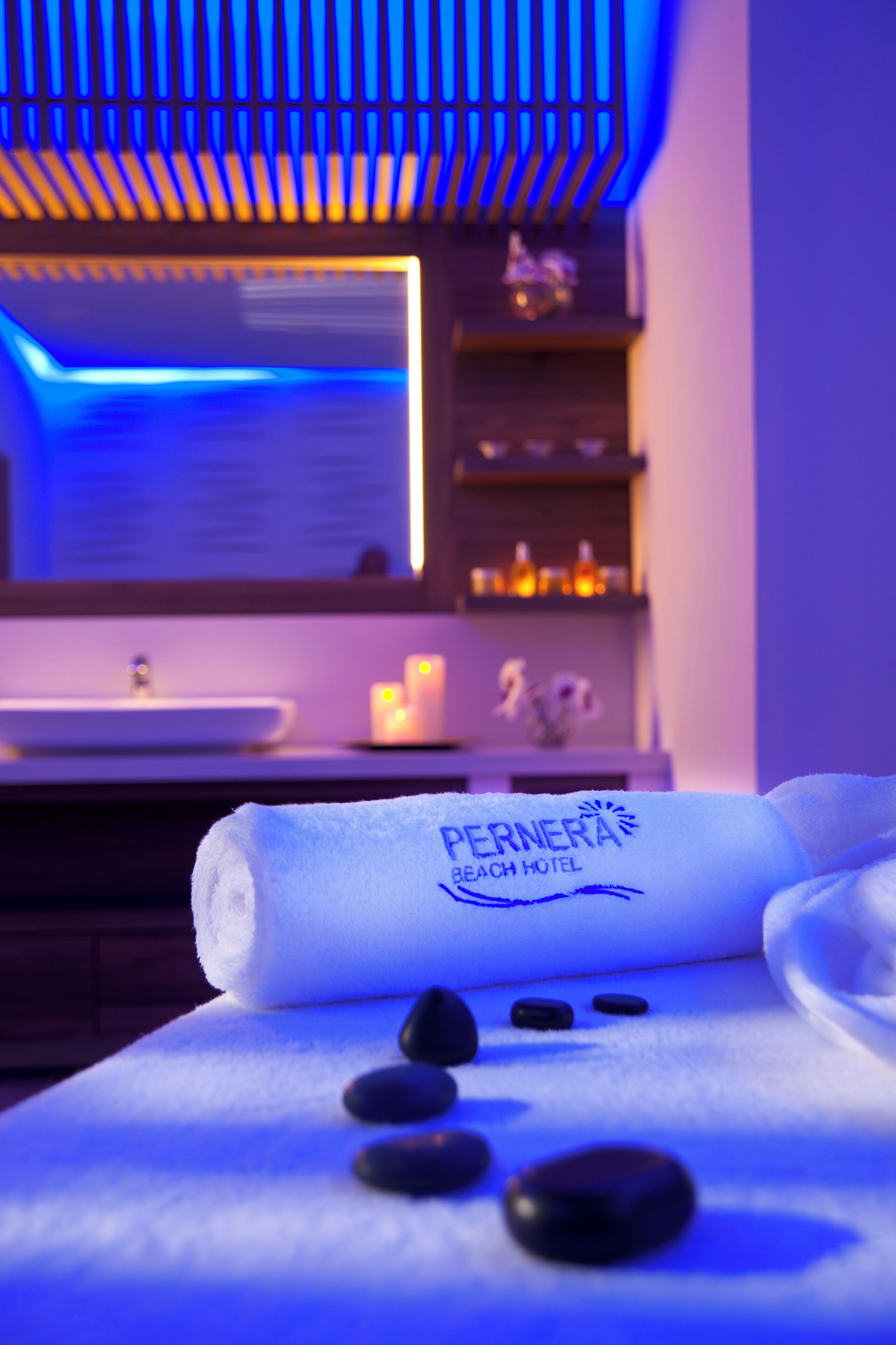 Una toalla con el logotipo de 'PERNERA BEACH HOTEL', un lavamanos, velas y una estantería con adornos.