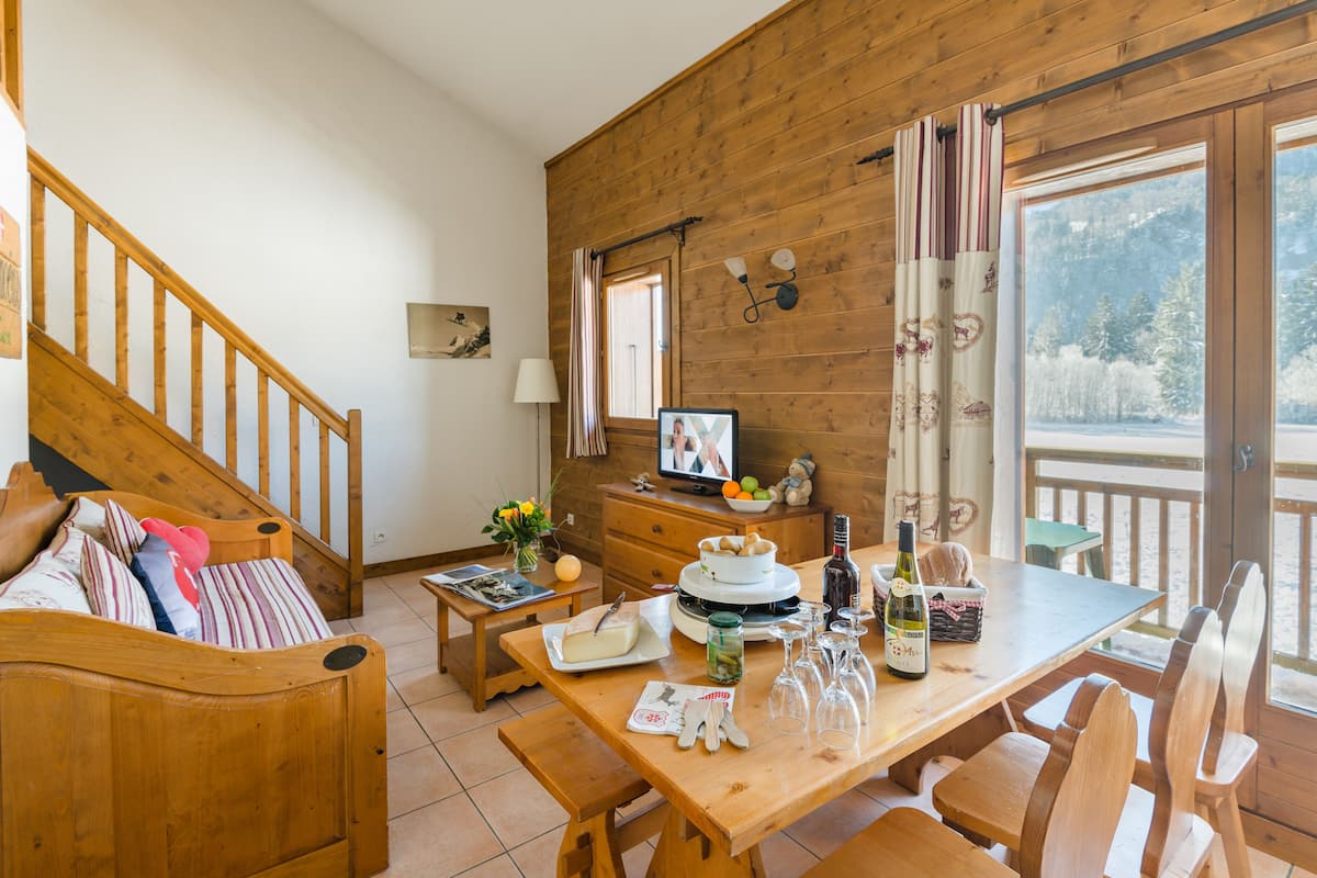 Een gezellig houten blokhutinterieur met een eettafel gedekt voor vier personen, een houten trap en een groot raam met uitzicht op een besneeerd landschap.