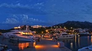 Un port de plaisance où sont amarrés plusieurs yachts, un bâtiment bien éclairé en arrière-plan et un paysage montagneux.