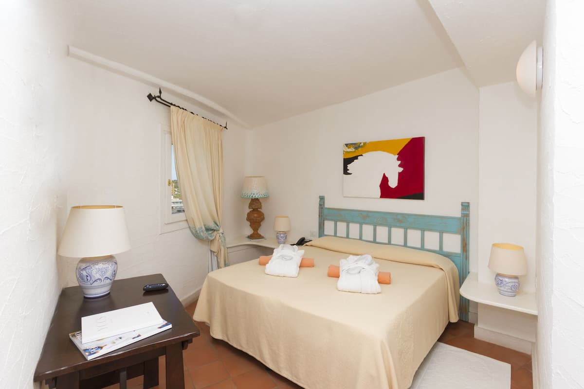 Een keurig opgemaakt slaapkamer met een bed, nachtlampjes, een nachtkastje, een ingelijst schilderij aan de muur, en een raam met gordijnen.