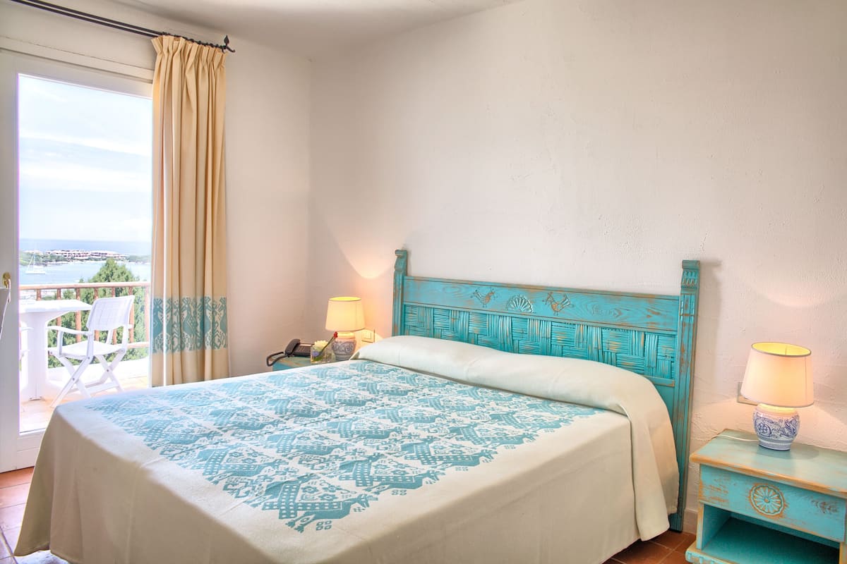 Een slaapkamer met een bed, nachtkastje, lamp, stoel en uitzicht op zee.