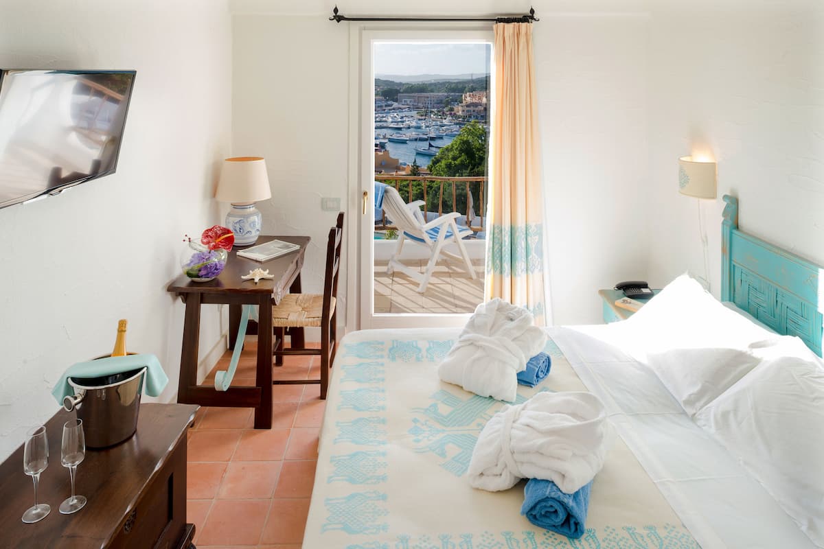 Hotelkamer met een bed, een bureau met champagne, een stoel en uitzicht op een jachthaven vanaf een balkon.