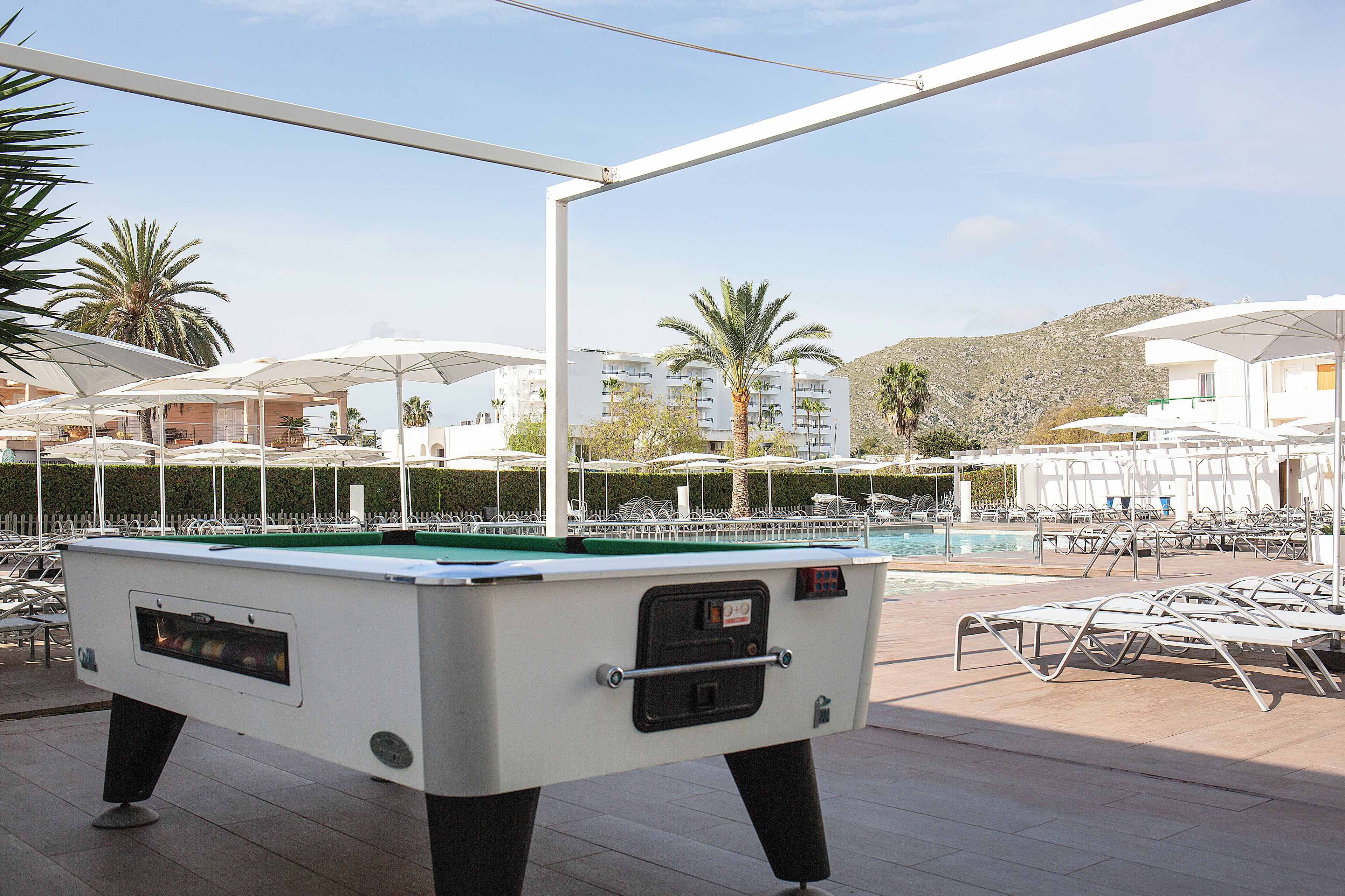 A pool table in an outdoor area with lounge chairs and umbrellas, surrounded by buildings and palm trees.