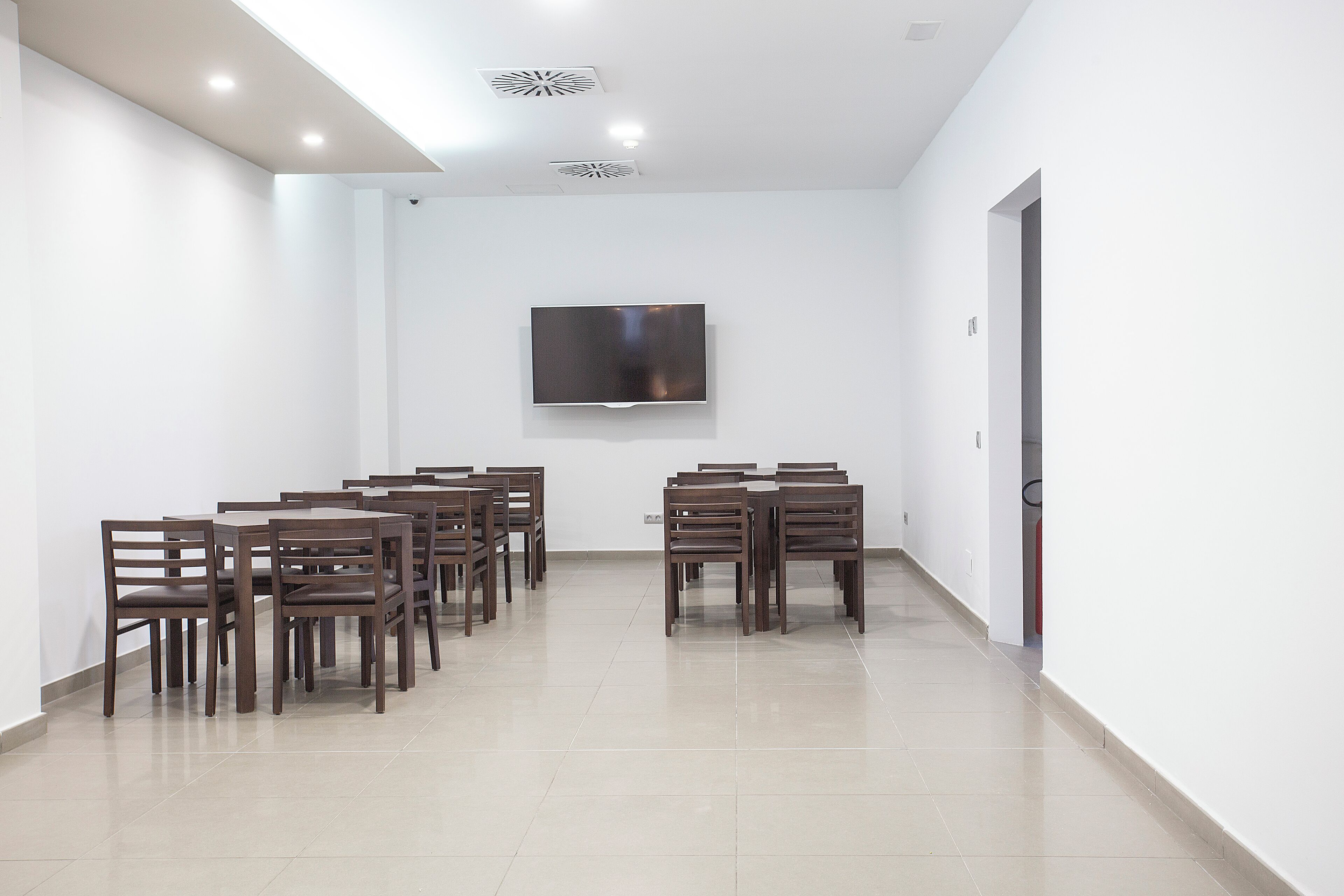A conference room with a flat-screen TV mounted on the wall, surrounded by wooden tables and chairs.