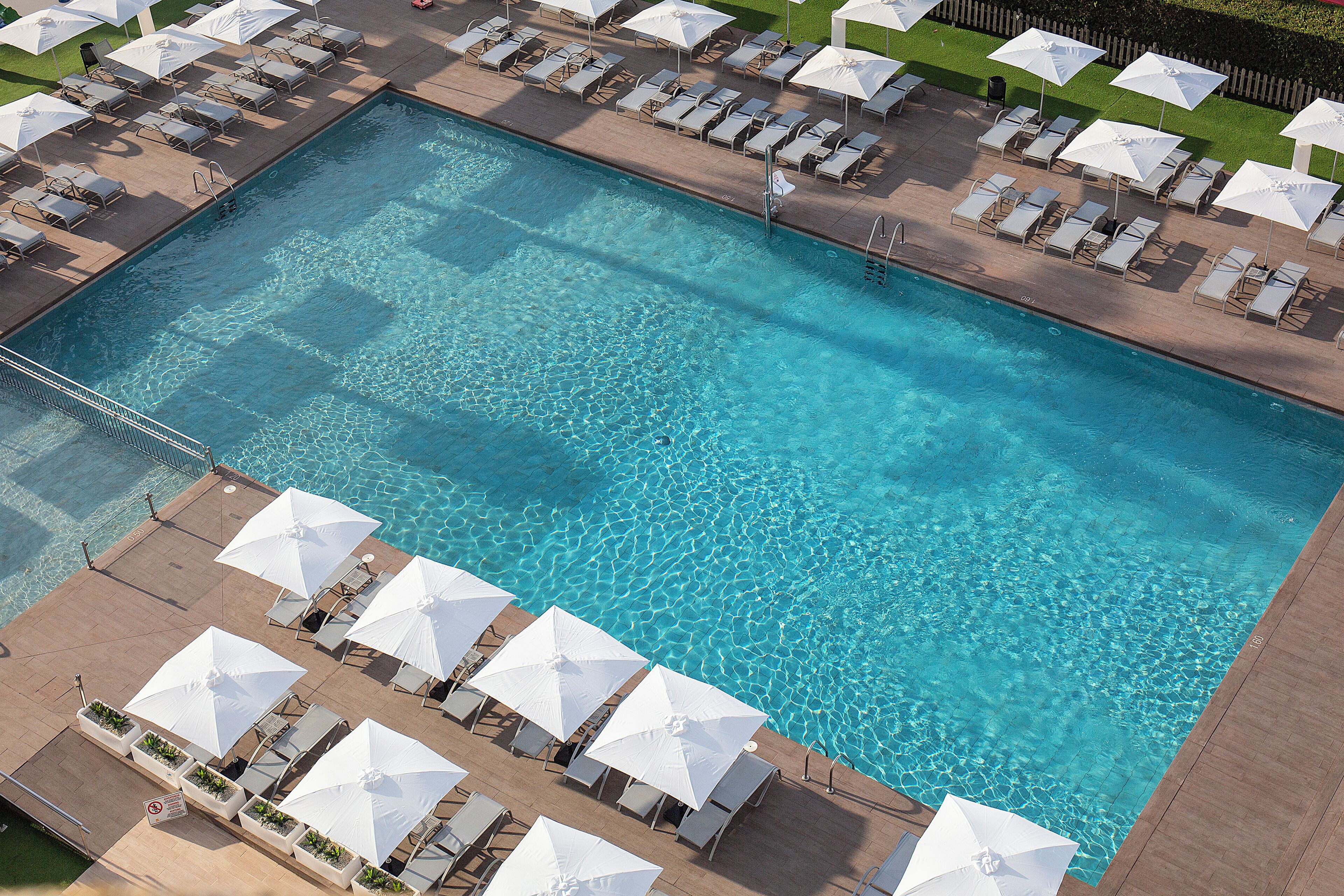 An aerial view of a rectangular swimming pool with clear blue water, surrounded by rows of white sun umbrellas and lounge chairs.