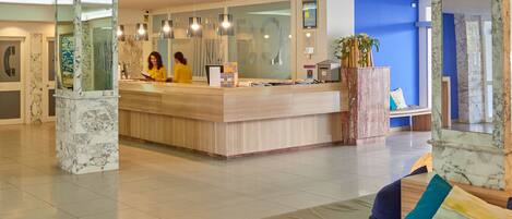 A hotel reception area with a marble reception desk, a seating area with a sofa and ottoman, and a large mirror reflecting the space.