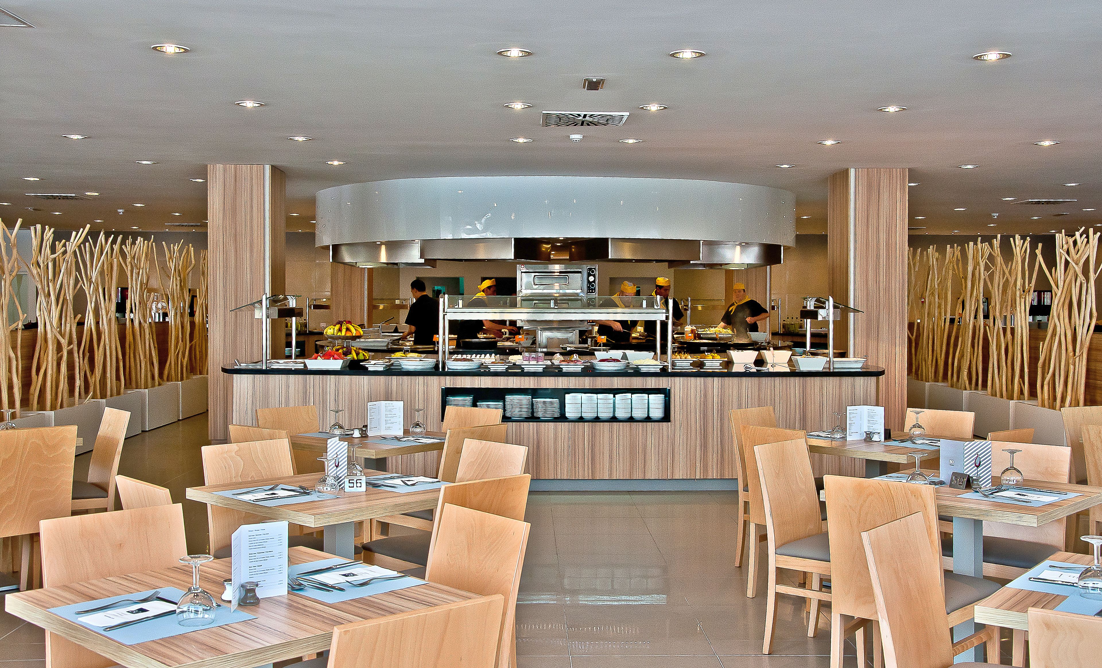 A modern restaurant with a central counter, multiple seating areas, and a high ceiling with recessed lighting.