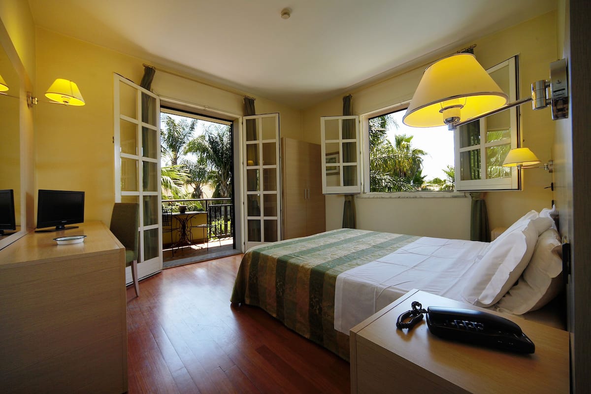 A hotel room with a bed, bedside table, lamp, TV, and a view of the outdoors.
