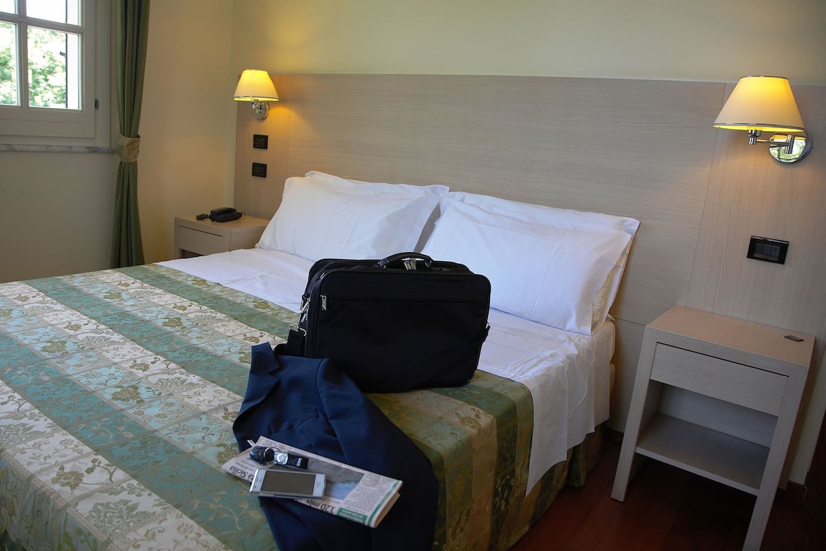 A hotel room with a bed, a bedside table, a lamp, and a suitcase on the bed.