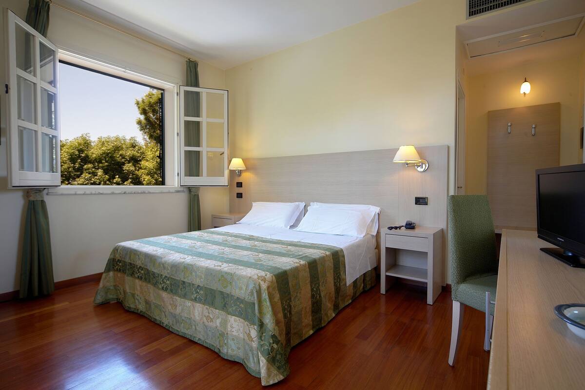 A hotel room with a bed, bedside table, lamp, chair, TV, and a window with curtains.