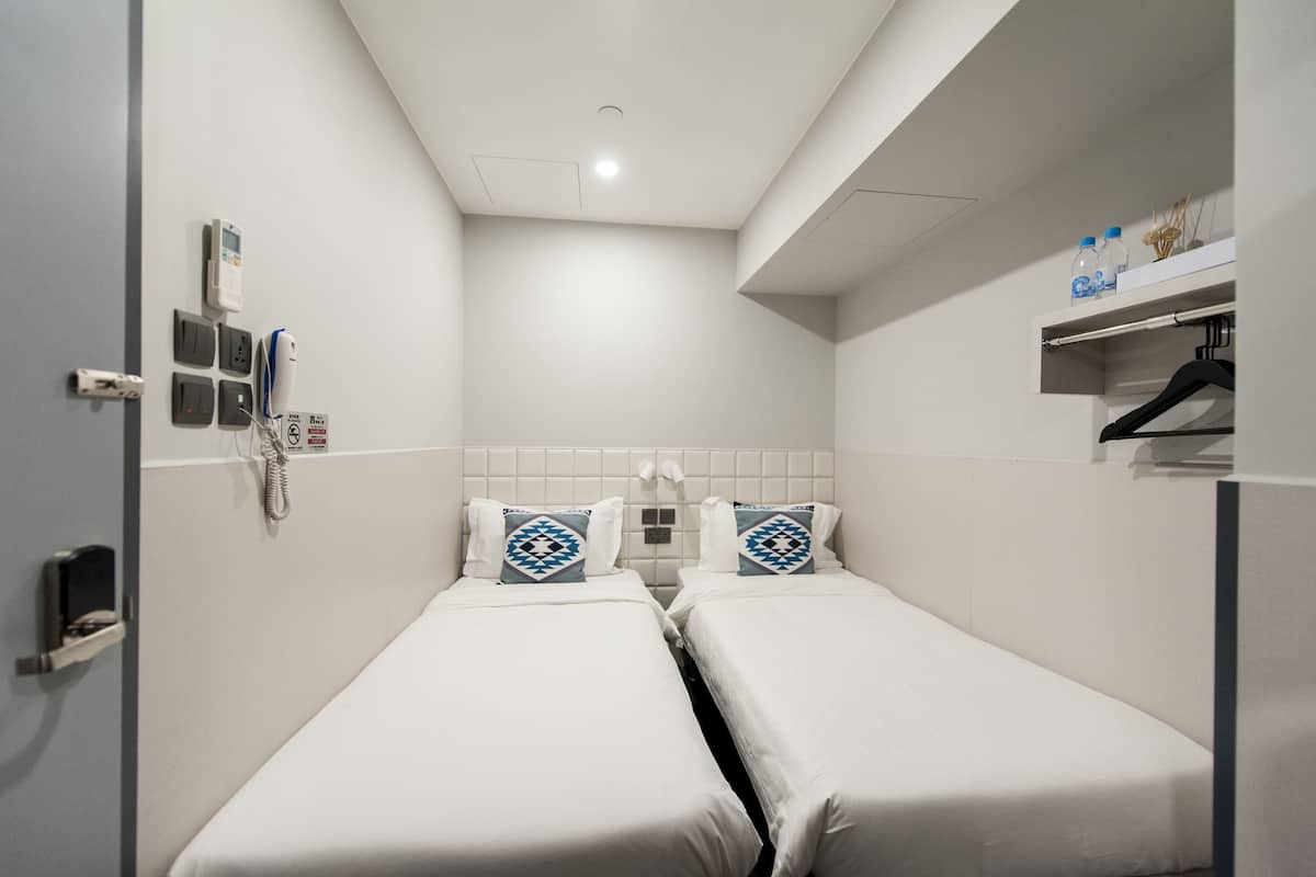 A small, clean room with two single beds, a bedside table, a shelf with personal items, and a wall-mounted phone.