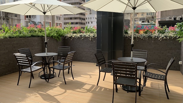 An outdoor patio with two round tables and four chairs, each with a white umbrella. The tables are set on a wooden deck, and the area is surrounded by a brick wall with a flower bed.