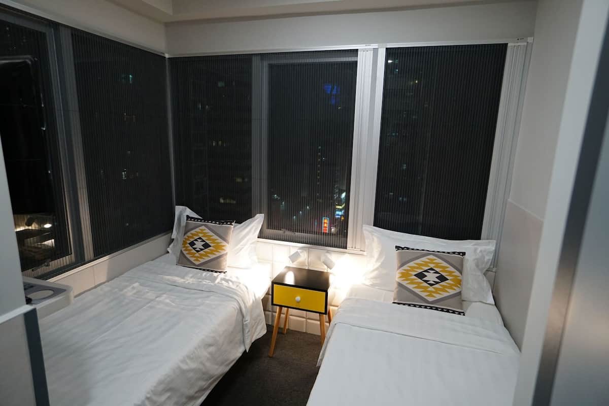 A hotel room with two beds, a small table, and large windows with blinds.
