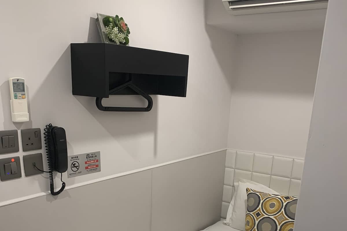 A small, neatly arranged hotel room with a single bed, a bedside table, a wall-mounted phone, and a small shelf with a plant.