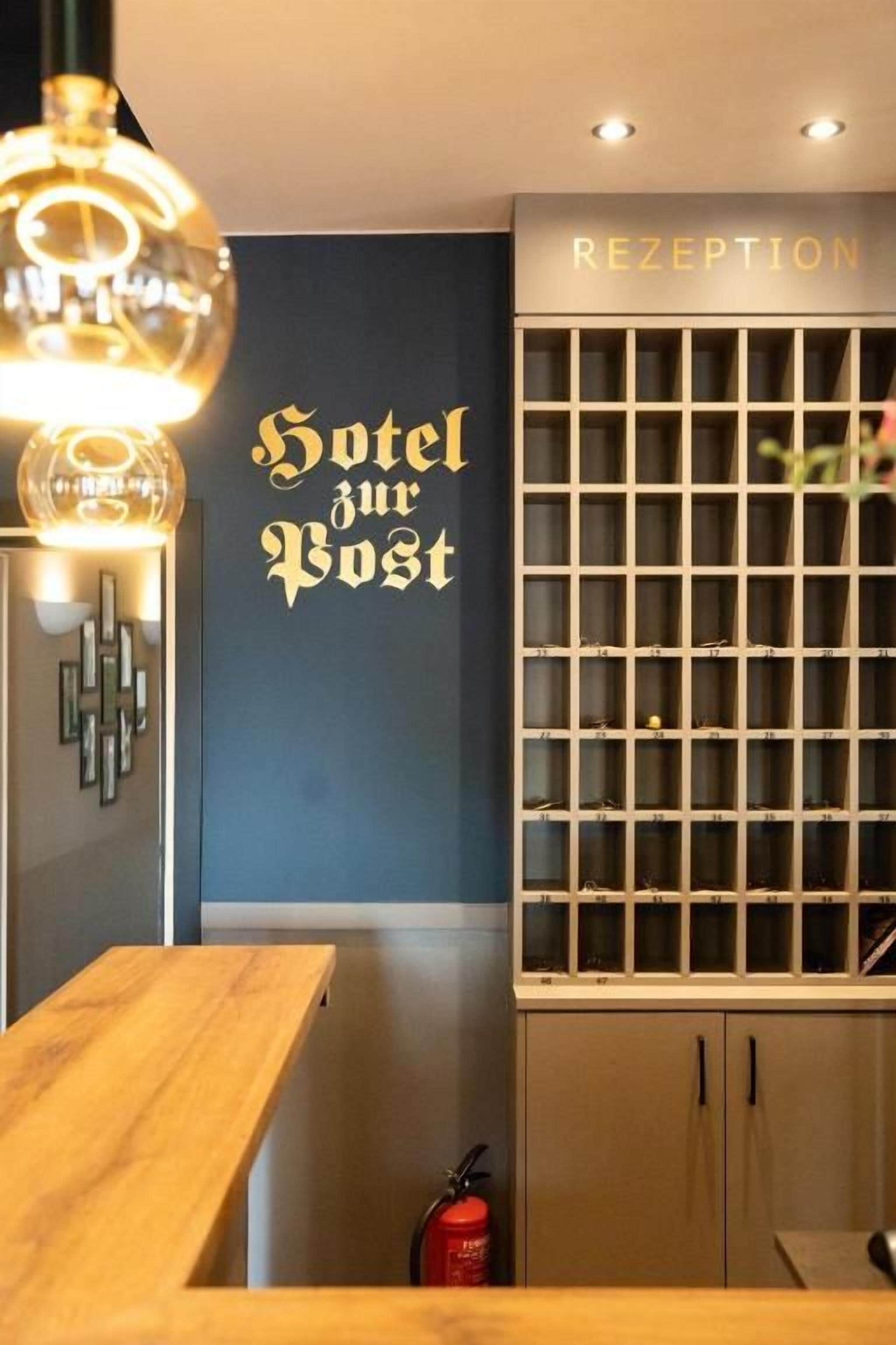 A reception area with a wall-mounted sign reading 'Hotel zur Post,' a reception desk, and a cabinet with drawers.