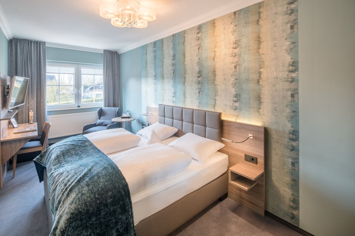 Quarto de hotel com uma cama grande, uma escrivaninha, uma cadeira, uma mesinha e vista para o exterior através de uma janela.
