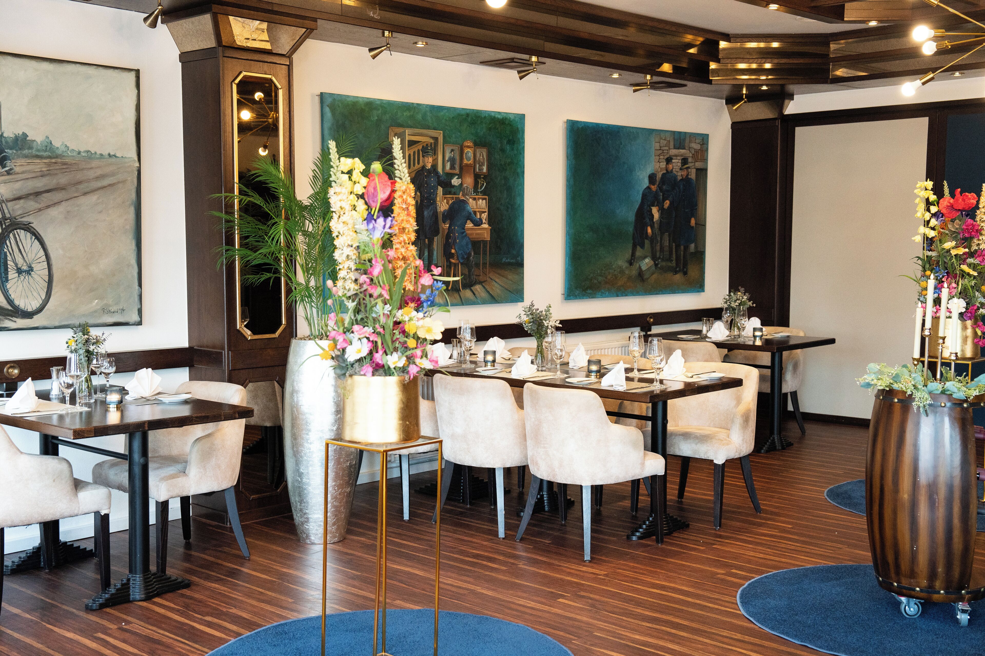 A restaurant with wooden flooring, round tables, and chairs. There are paintings on the walls and a large vase with flowers on the tables.