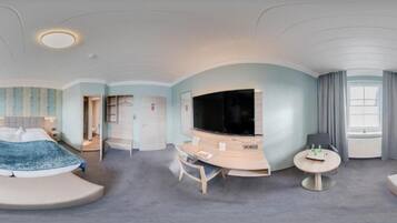 A modern hotel room with a curved bed, a desk, a flat-screen TV, and a seating area.