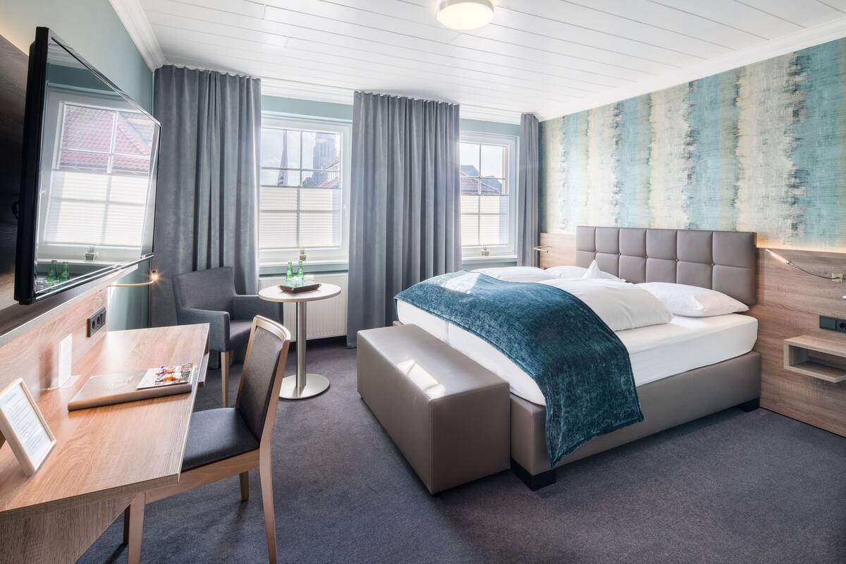 Quarto de hotel moderno com uma cama grande, uma escrivaninha e uma cadeira. Há duas janelas com cortinas, uma mesinha e uma parede com design texturizado.