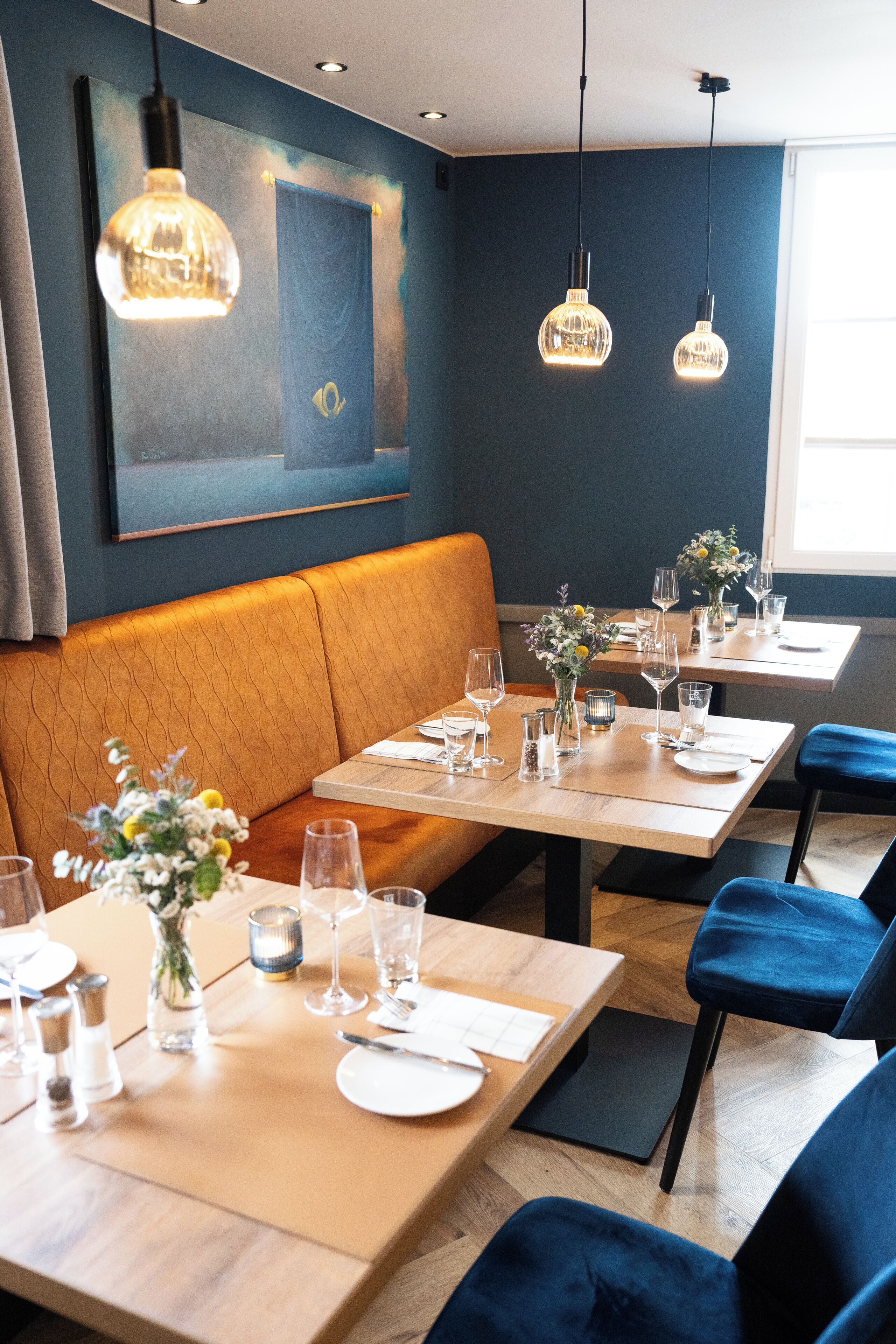 A restaurant with a modern interior, featuring wooden tables, blue upholstered chairs, and pendant lighting.