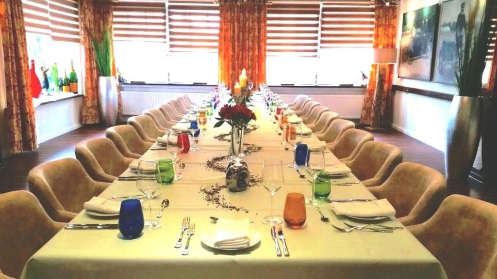 Una larga mesa de comedor preparada para una comida, con sillas a ambos lados, un jarrón con flores en el centro y botellas de vino en el alféizar de la ventana.
