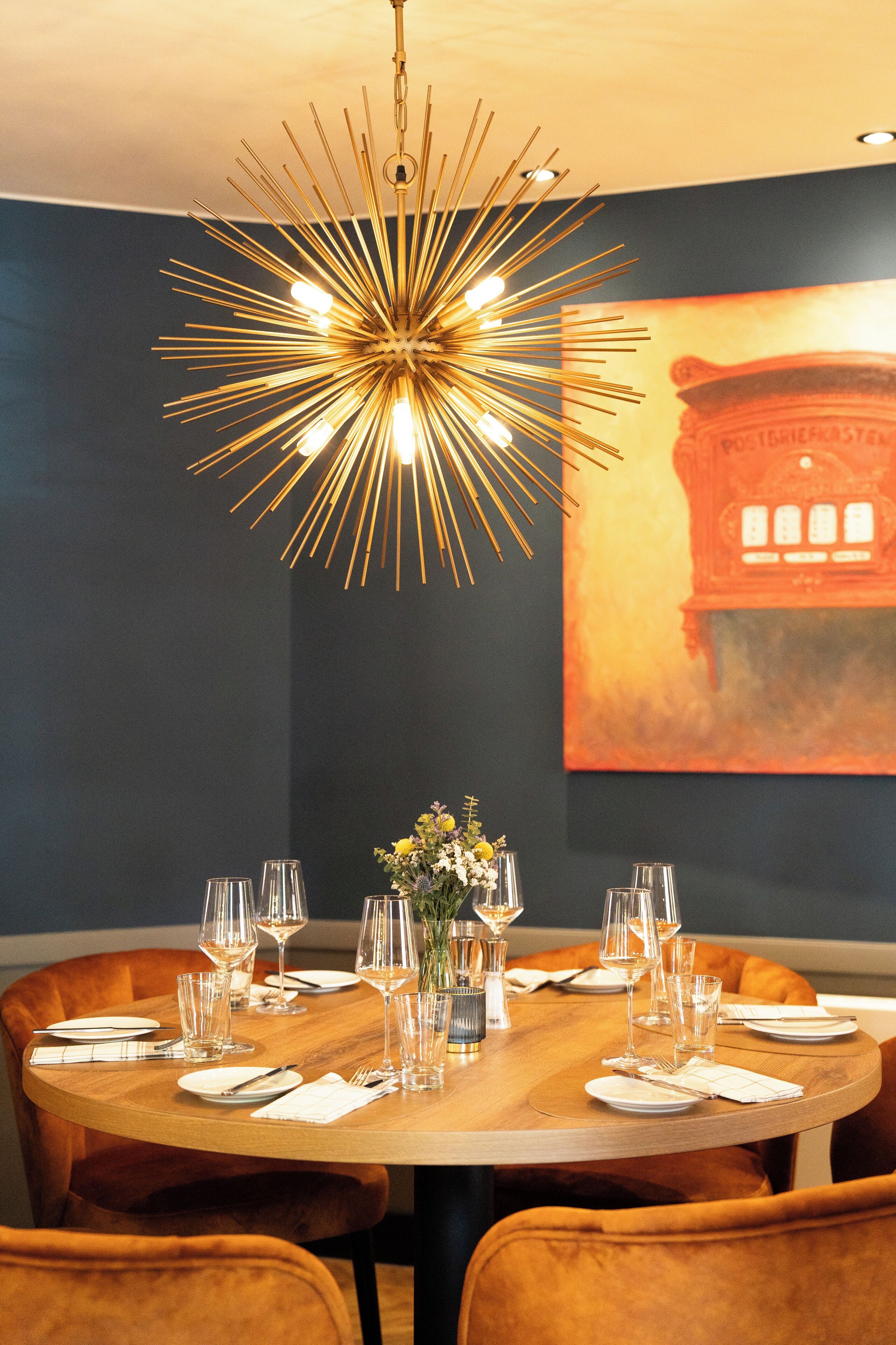 A dining area with a round table set for six, a modern chandelier, and a large abstract painting on the wall.