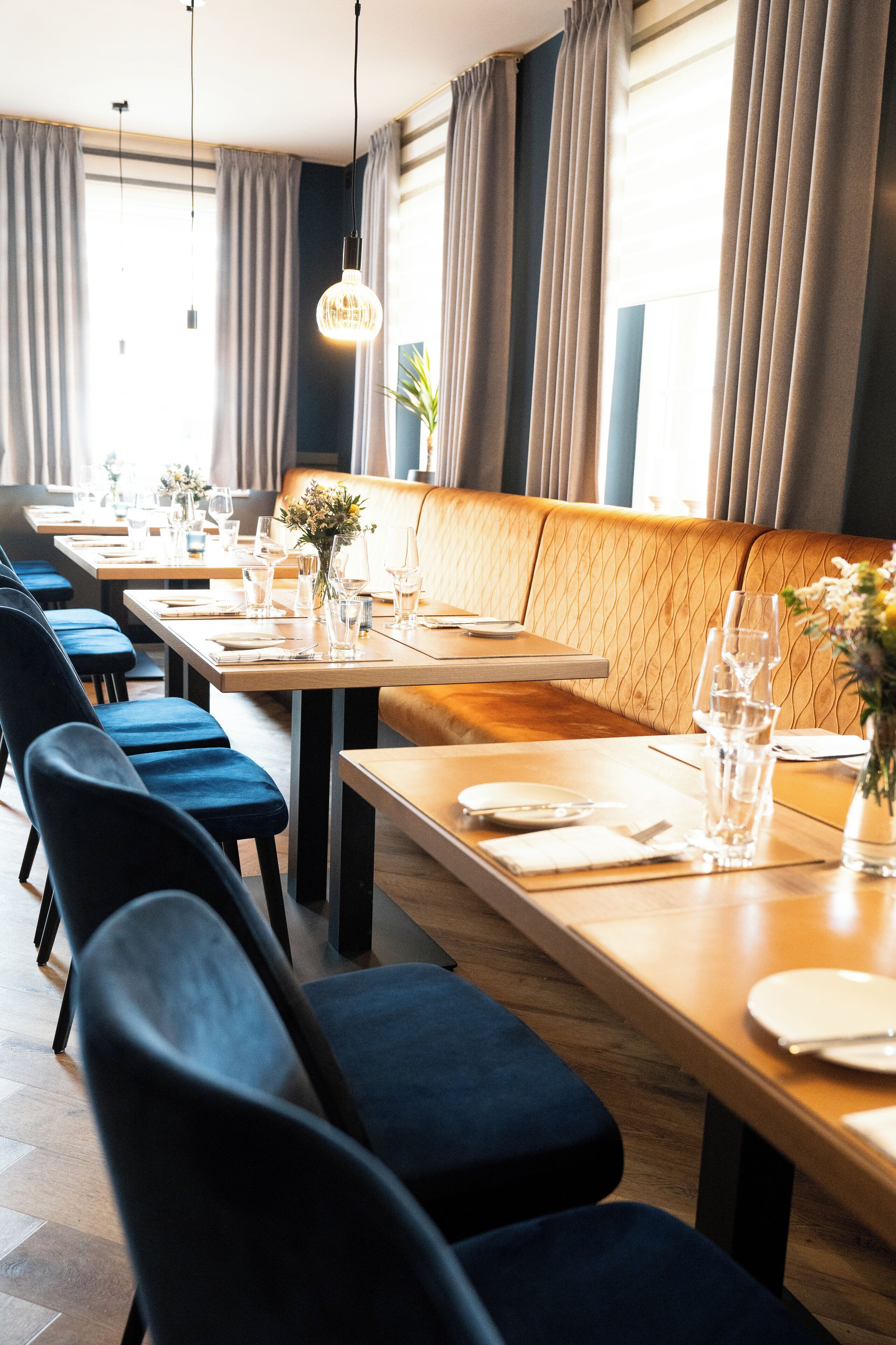 A restaurant with long wooden tables, blue upholstered chairs, and a cushioned bench.