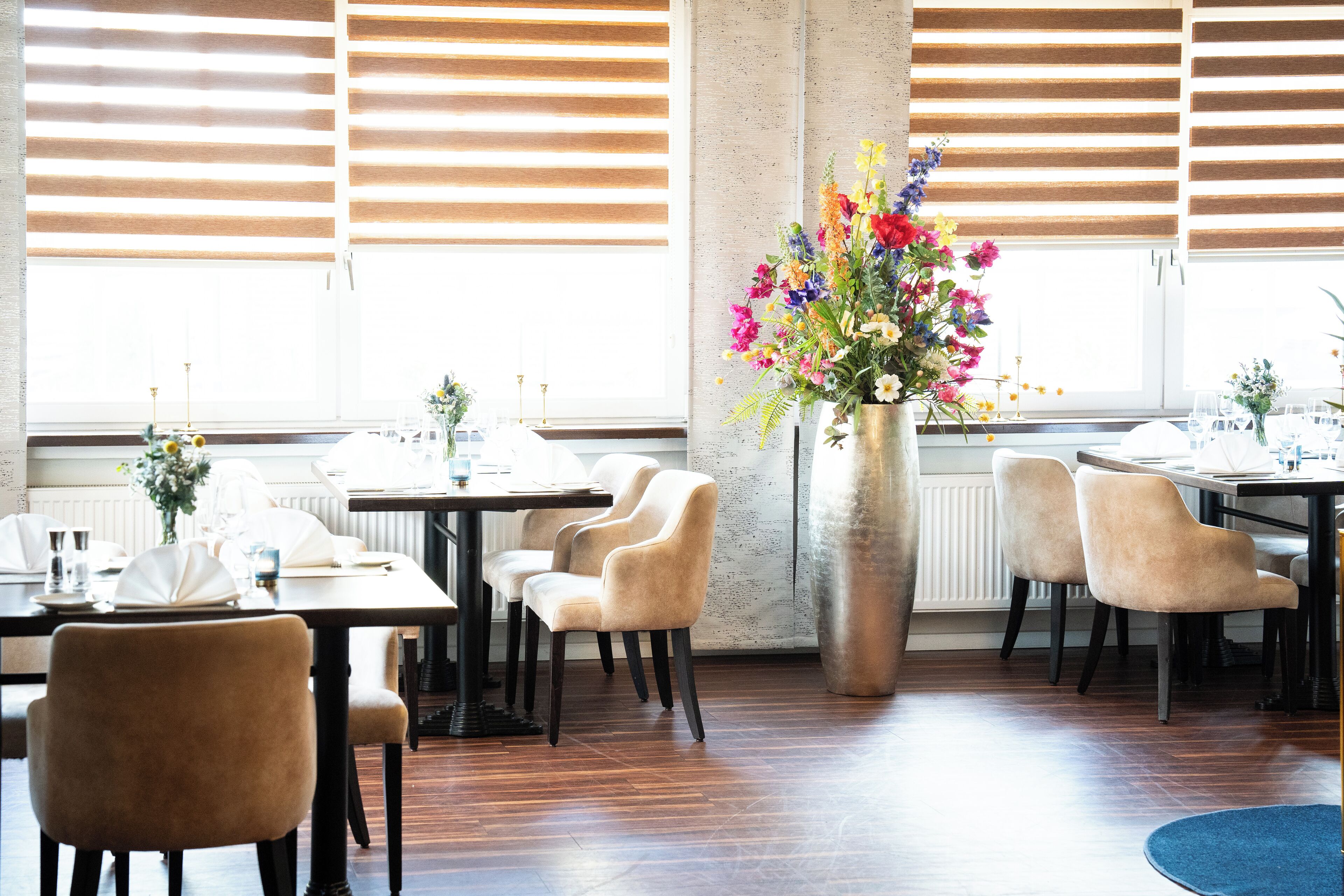 A dining area with wooden tables and chairs, a large vase of flowers, and large windows with blinds.