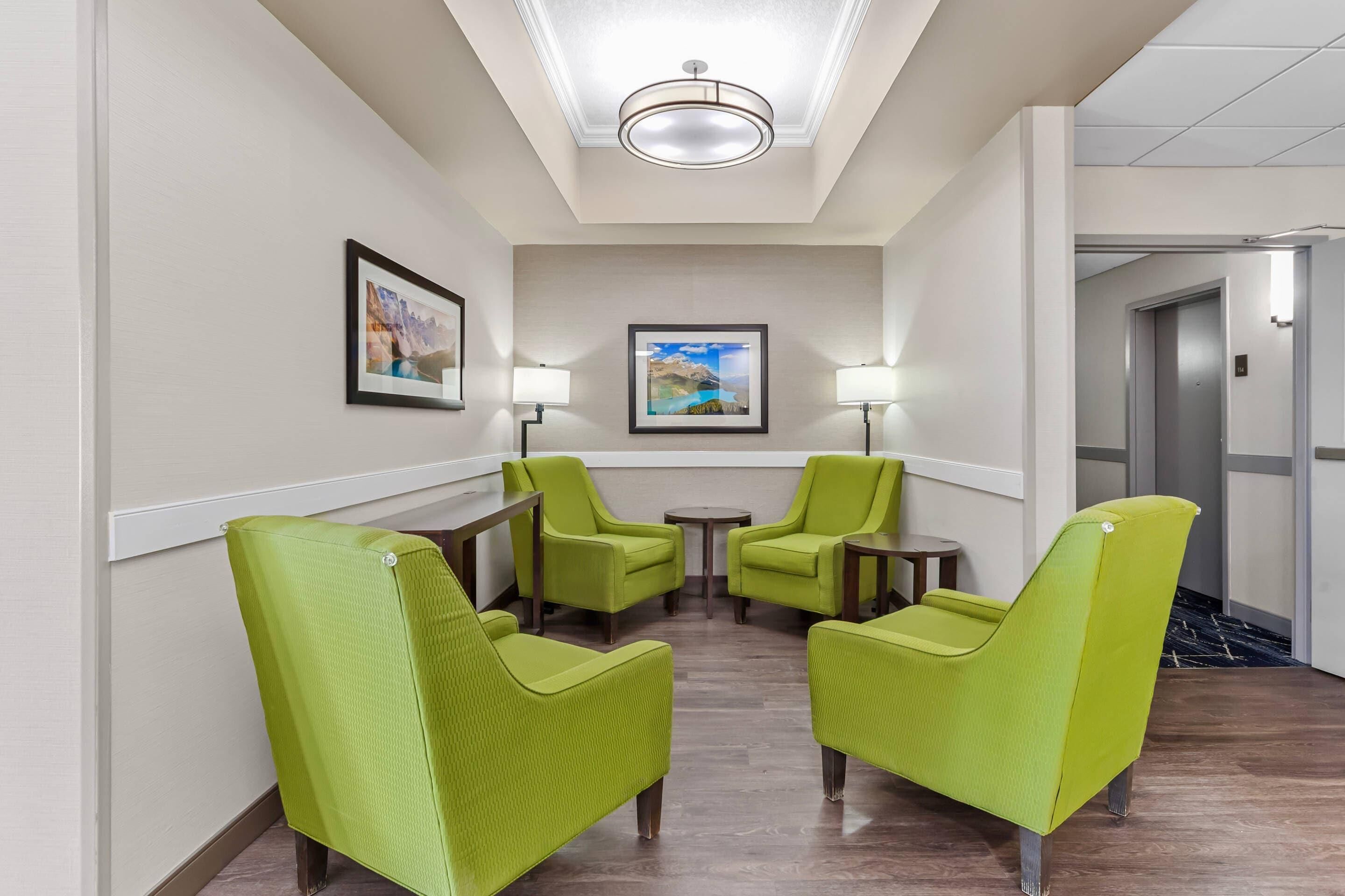 Um hall de hotel com uma mesa de recepção, dois poltronas verdes, uma mesinha, duas luminárias de chão e um quadro na parede.