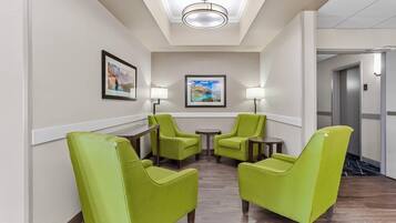 A hotel lobby with a reception desk, two green armchairs, a small table, two floor lamps, and a framed picture on the wall.