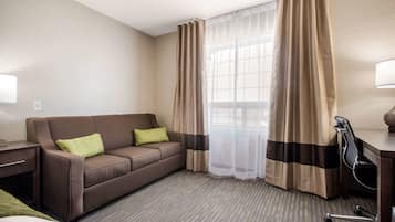 A hotel room with a brown sofa, a desk, a chair, and a window with sheer curtains.