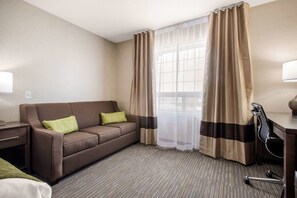 Une chambre d’hôtel avec un canapé marron, un bureau, une chaise et une fenêtre ornée de rideaux légers.