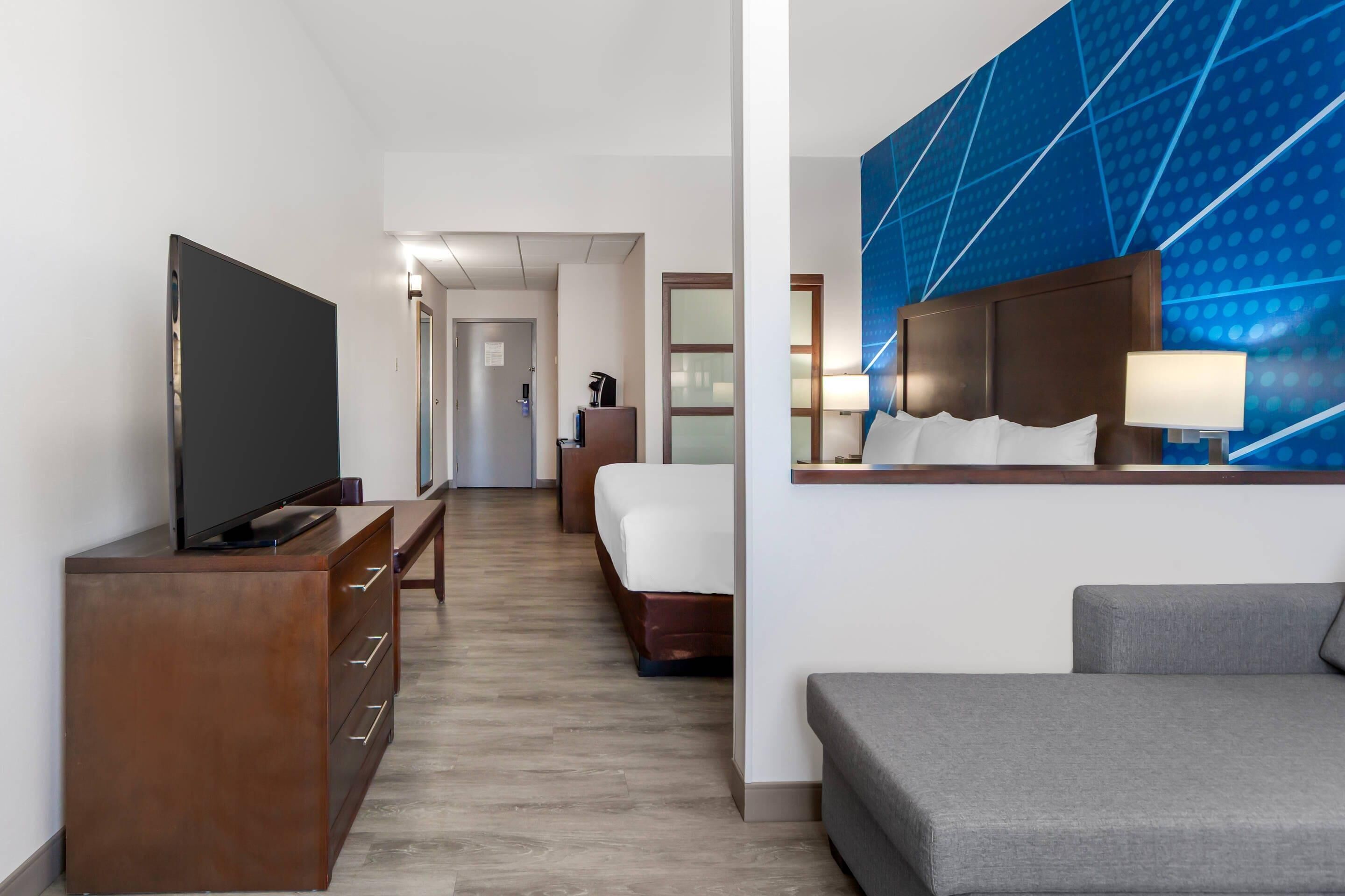 A hotel room with a flat-screen TV, a wooden dresser, a bed, a gray sofa, and a blue patterned wall.