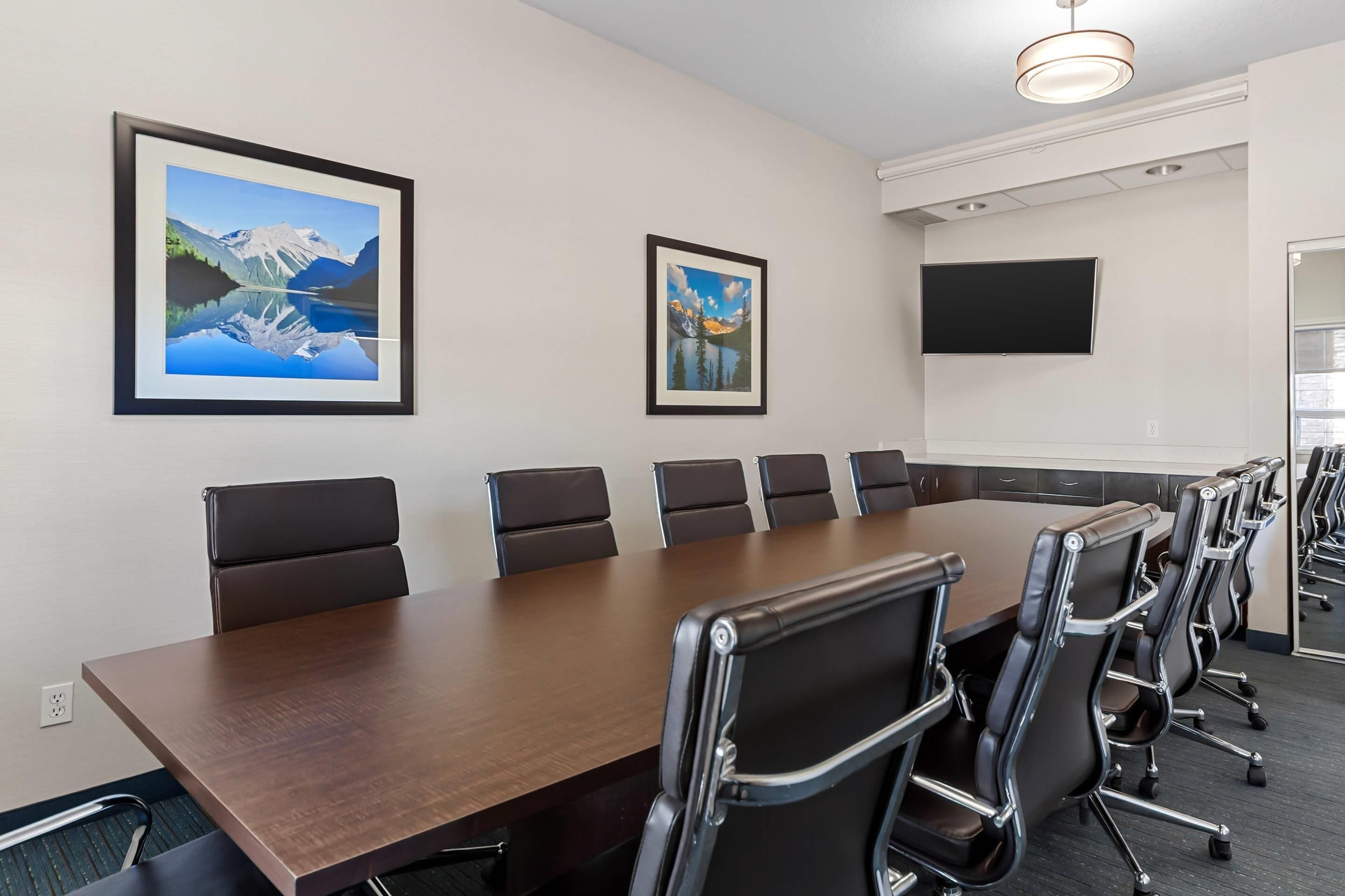 Una sala de conferències amb una taula llarga, sis cadires, dos quadres de paisatges amb marc, un televisor de pantalla plana i una làmpada al sostre.