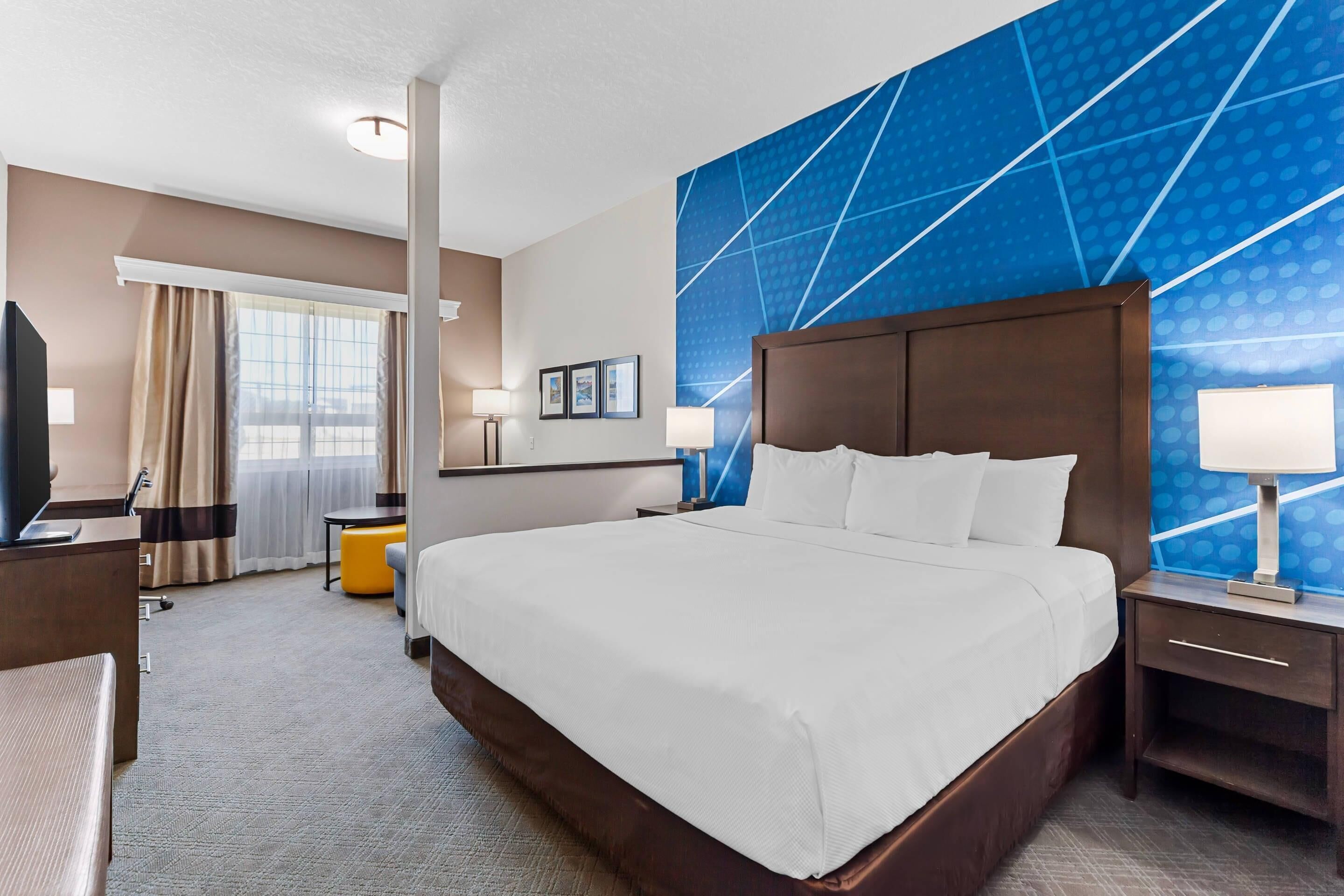 Ein Hotelzimmer mit einem großen Bett, einem Schreibtisch mit Computer, einem Stuhl und einem Fenster mit Vorhängen.