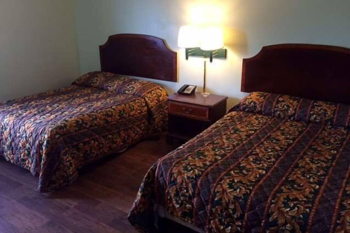 A room with two beds, each with a patterned bedspread and a wooden headboard. There is a nightstand with a lamp between the beds.