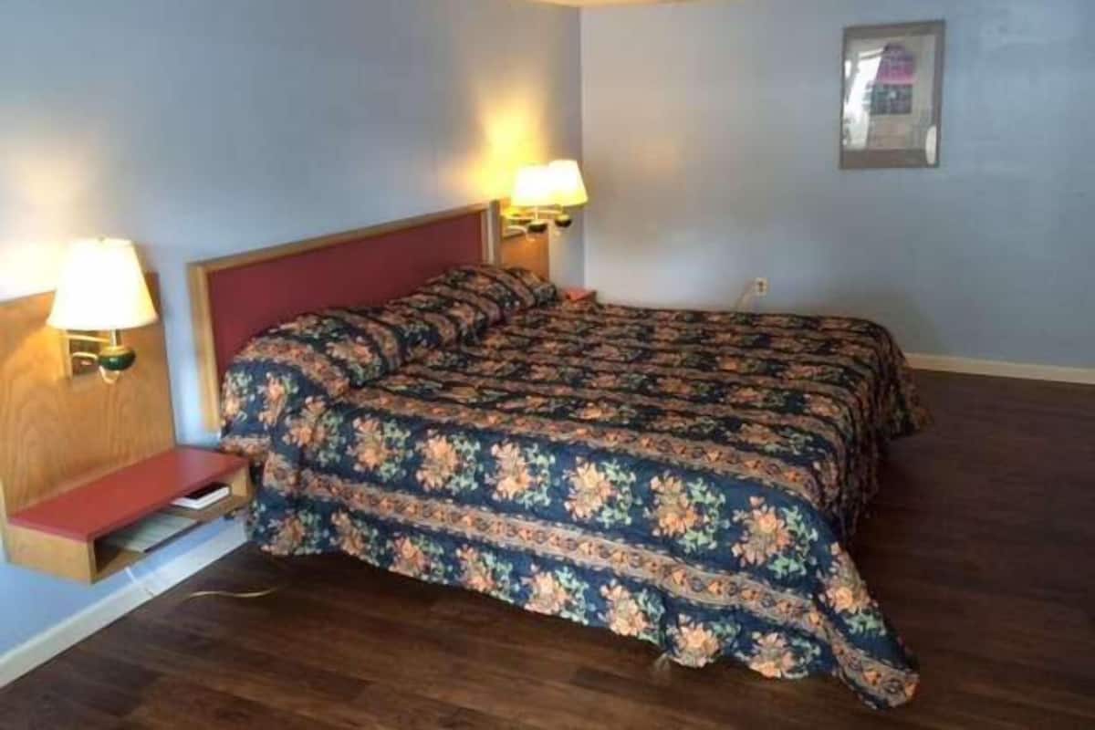 A hotel room with a double bed, two bedside lamps, a small wooden nightstand, and a framed picture on the wall.