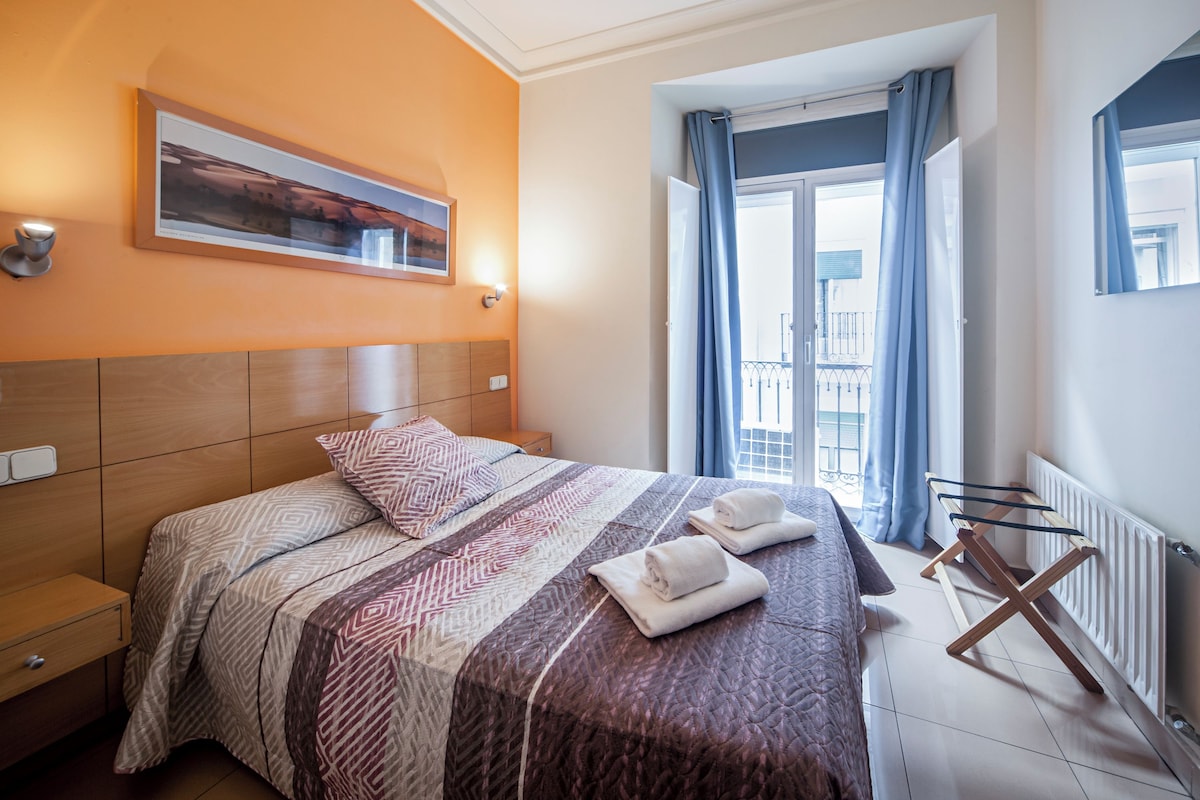 A hotel room with a large bed, a headboard, a window with curtains, a radiator, and a wall-mounted picture.