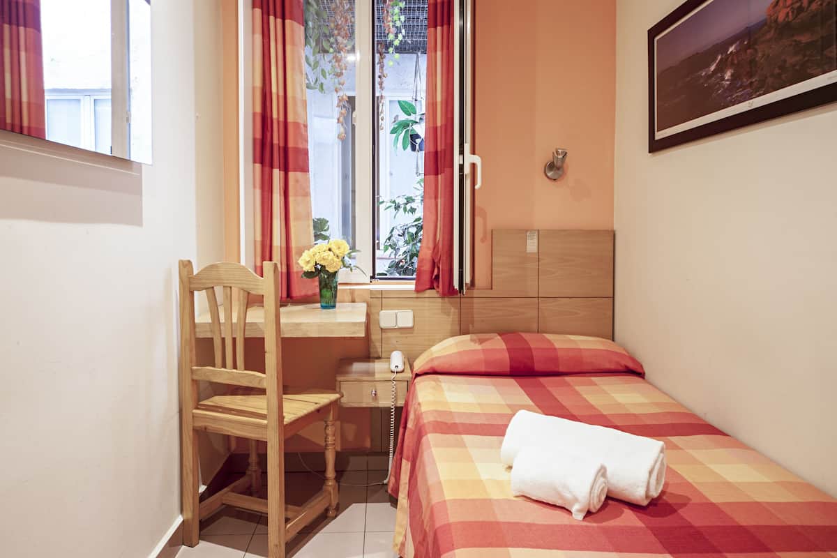 A small, tidy room with a bed, a desk, and a chair. There is a window with curtains, a plant on the desk, and a painting on the wall.