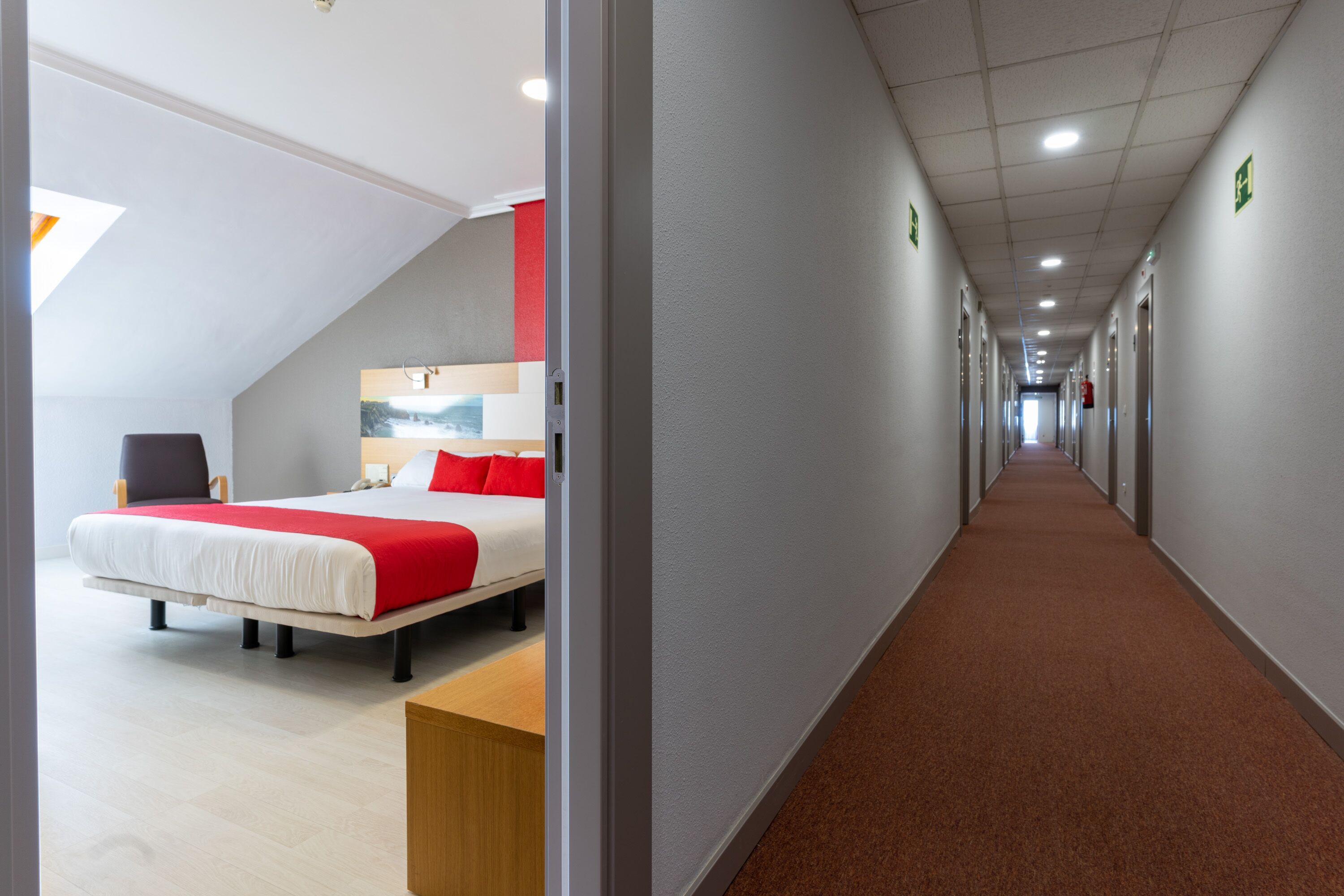 A hotel corridor with a long hallway, a bed with red bedding, and a wooden bench.