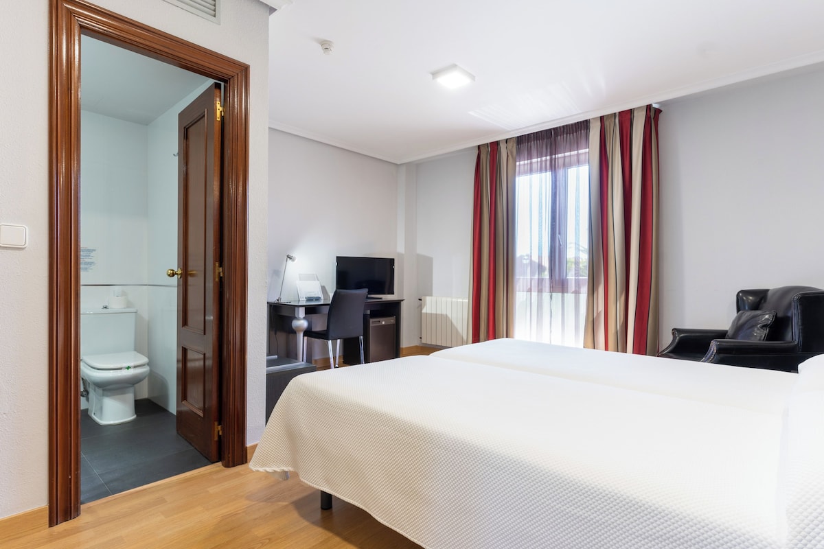 Une chambre d’hôtel équipée d’un lit, d’un bureau, d’une chaise et offrant une vue sur l’extérieur grâce à une fenêtre avec des rideaux.