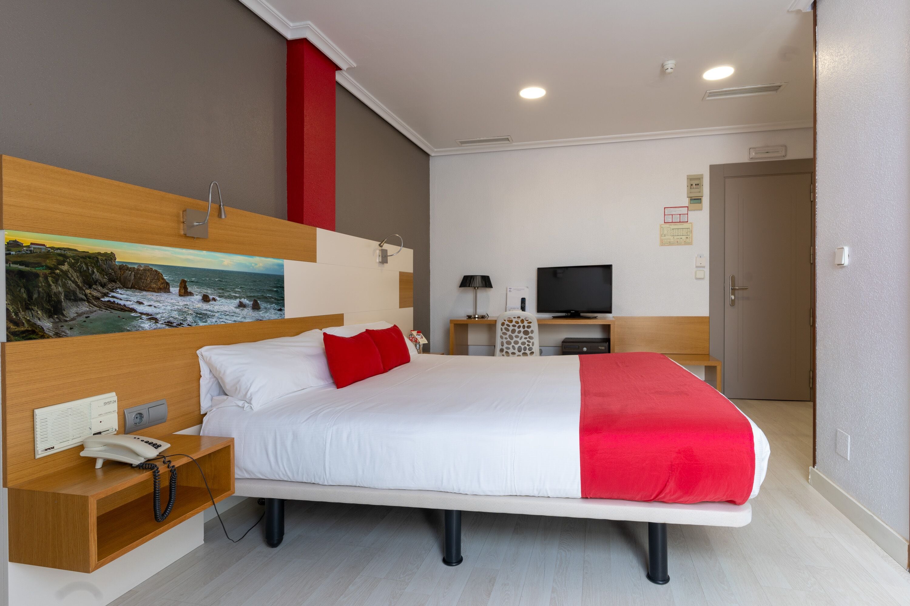 A hotel room with a large bed, a wooden headboard, a painting of a coastal scene, a desk with a lamp, and a television.