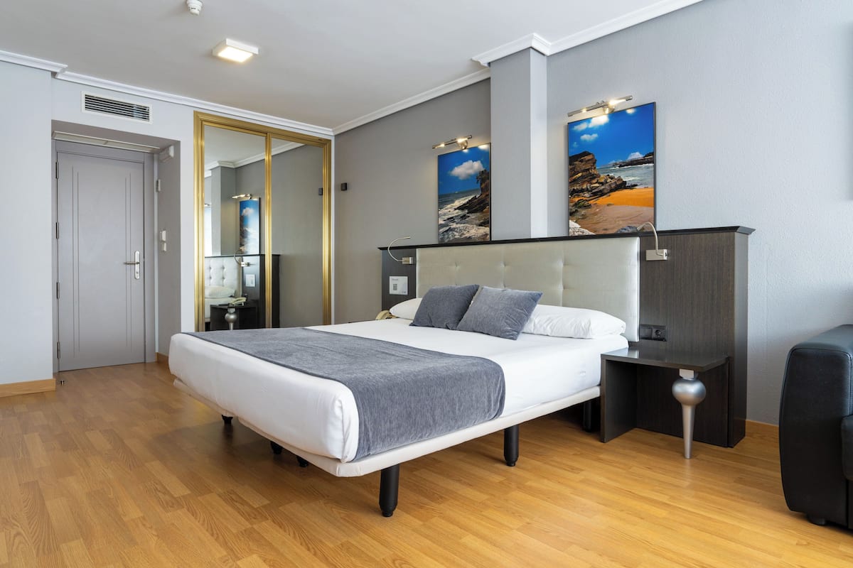 A modern hotel room with a large bed, a bedside table, a nightstand, a mirror, and two wall-mounted paintings.