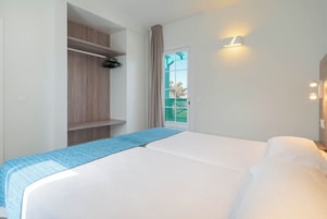 A hotel room with a bed, a wooden wardrobe, a window with curtains, and a wall-mounted light fixture.