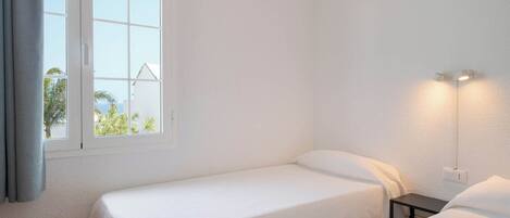Ein Zimmer mit zwei Einzelbetten, einem fenster mit Vorhängen, einer Wandlampe und Blick auf Palmen.